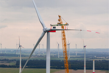 A large crane installs a rotor blade on a wind turbine. Numerous other turbines stand in the wide landscape in the background.