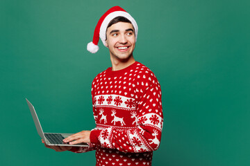 Merry young IT man wear red warm Christmas sweater Santa Claus hat posing hold use work on laptop...