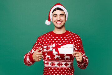 Merry young man wear red Christmas sweater Santa Claus hat posing hold gift coupon voucher card for...