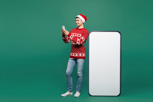 Full body merry young man in red Christmas sweater Santa Claus hat posing big huge blank screen area use mobile cell phone smartphone device isolated on plain green background Happy New Year concept