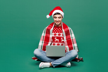 Full body merry young fun IT man wear warm cozy plaid red Santa Claus hat posing sit hold use work...