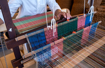 Doha, Qatar - December 01, 2025: Traditional hand-weaving in progress, showing a close-up view of colorful threads arranged on a wooden loom. A craftsman&rsquo;s hands guide the weaving process.