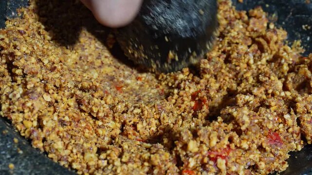 Hand grinding peanut chili paste using a traditional stone mortar. Fresh spices combine with roasted nuts to create authentic Indonesian cooking ingredient