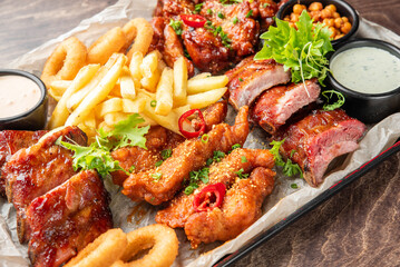 A vibrant, close-up shot of an assorted comfort food platter featuring BBQ pork ribs, crispy fried chicken strips, golden french fries, and onion rings with dipping sauces