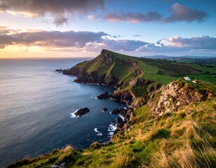 Coastal headland covered in green grass meets the sea under a cloudy, colorful sunset sky