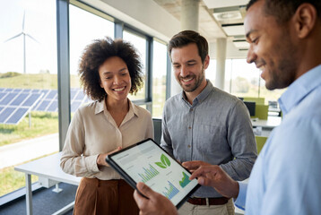 Colleagues reviewing sustainability analytics on digital tablet in modern office with solar panels and wind turbine outside, clean energy strategy and corporate environmental reporting teamwork