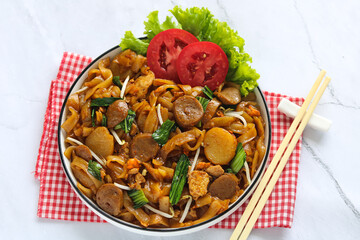 Stir-Fried Flat Rice Noodles. A savory dish featuring wide rice noodles with sliced meatballs, fish cakes, egg, greens, and bean sprouts. On the side is a pair of chopsticks.