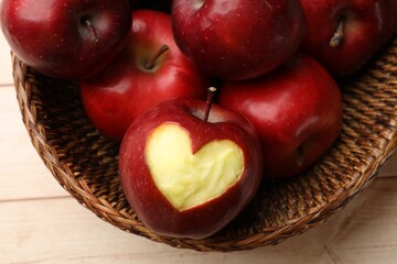 Ripe apple with carved heart and whole ones in wicker basket on light wooden table, top view