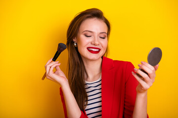 Charming woman in red jacket smiling while applying makeup with a brush, cheerful image on a bright...
