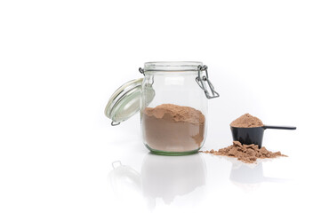 Horizontal image of a transparent jar of chocolate whey protein next to a scoop. White background with a reflective surface.