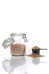 Vertical image of a transparent jar of chocolate whey protein next to a scoop. White background with a reflective surface.