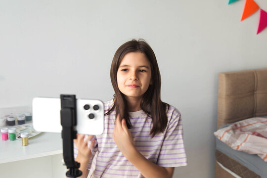 young girl recording a vlog on her smartphone at home, adjusting hair while creating content. Bright, cozy bedroom setting with creative supplies, showing modern child vlogging  digital expression