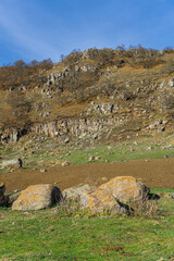 Obraz premium Huge rocks in a clearing with autumn-colored green and yellow grass. The cliff is covered with leafless bushes and trees. A bright blue sky is in the background.