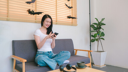 Young woman smiling while using smartphone at home with cozy interior, decorative bats on window and indoor plant in bright lighting