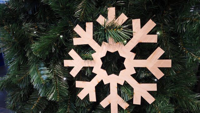 Wooden snowflake ornament hangs on a green Christmas tree with small lights peeking through the branches. The scene conveys a festive winter atmosphere and holiday decoration concept