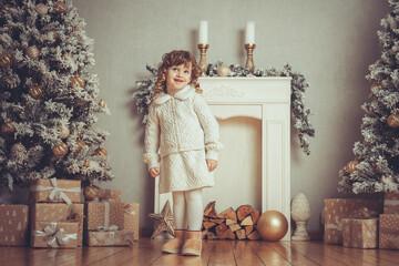 H&uuml;bsches kleines M&auml;dchen mit Locken steht vor einem Kamin neben einem Weihnachtsbaum zu Weihnachten bei Abendstimmung mit Lichtern, Var. 1