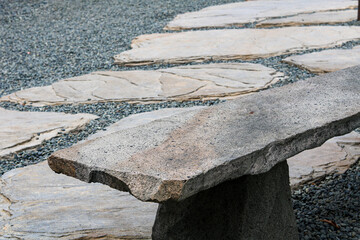 A view of a natural stone bench, crafted from rough-cut slab, overlooking a tranquil garden path of flagstones and dark grey gravel.