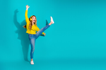 Young woman in yellow sweater and jeans jumps with headphones against blue background showing...