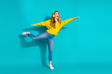 Young woman in yellow sweater wearing headphones jumps in a bright blue studio for a joyful fashion...