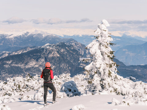A woman with a backpack snowshoes on a snowy mountain, looking at the view. Paganella, Roda refuge,Trentino, Italy