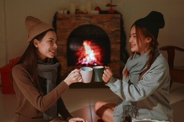 Fototapeta premium Teen girls having fun together on holiday, sitting in front of the fireplace. Asian lesbian couple celebrating Christmas, drinking warm drinks to keep warm.