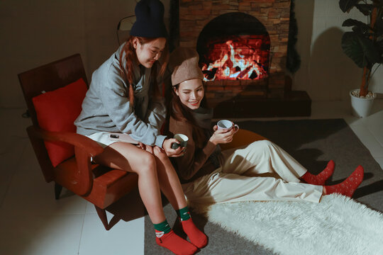 Teen girls having fun together on holiday, sitting in front of the fireplace. Asian lesbian couple celebrating Christmas, drinking warm drinks to keep warm. - Powered by Adobe
