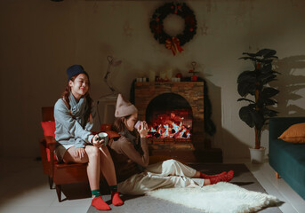 Obraz premium Teen girls having fun together on holiday, sitting in front of the fireplace. Asian lesbian couple celebrating Christmas, drinking warm drinks to keep warm.