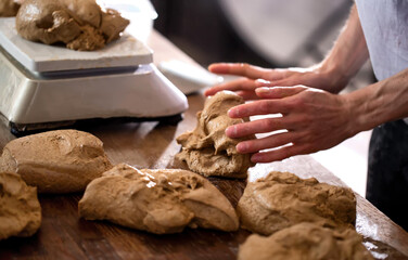 Close up view of hands shaping fresh bread dough on a wooden surface, ready for baking. Preparation of artisan food with multiple dough portions and a scale visible in bakery or kitchen environment.