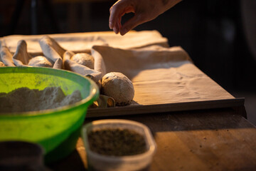 Hand of baker preparing dough rolls on baking paper for fresh bread baking, flour in green bowl and spice container visible on rustic wooden table, dark background