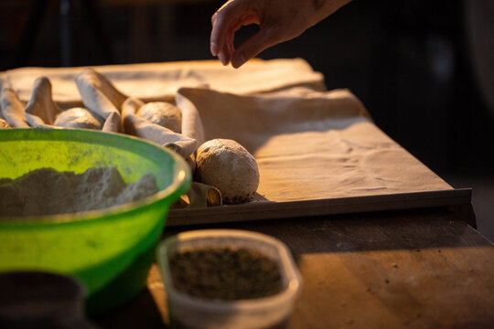 Hand of baker preparing dough rolls on baking paper for fresh bread baking, flour in green bowl and spice container visible on rustic wooden table, dark background - Powered by Adobe