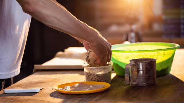 Hands of baker preparing dough, dipping round bread roll in seeds on rustic wooden table, bathed in warm golden light of golden hour, process of artisanal baking creation