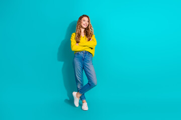 Young woman in yellow cardigan and jeans stands against blue backdrop smiling confident and relaxed
