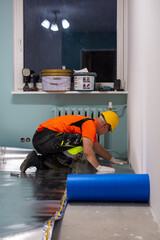 A finishing worker applies an insulating underlayment with an aluminum layer under wooden floor panels. Sound and thermal insulation. Apartment renovation.