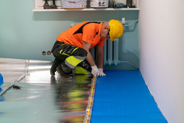 A finishing worker applies an insulating underlayment with an aluminum layer under wooden floor panels. Sound and thermal insulation. Apartment renovation.