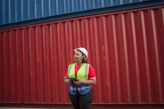 Black female logistics worker wearing a safety vest and helmet holding tablet while standing and looking with vision in a shipping container yard, show digital cargo management and modern logistics.