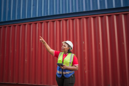 Black female logistics worker wearing a safety vest and helmet holding tablet while point in the air in shipping container yard, representing digital cargo management and modern logistics technology. - Powered by Adobe