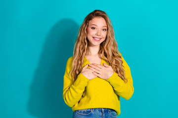 Bright young woman in a yellow sweater smiles with hands over heart against blue background