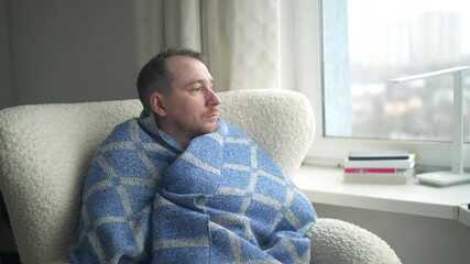 A man sits peacefully by a window, wrapped in a warm blanket. He gazes outside, deep in thought, while savoring a soothing drink on a cold day.