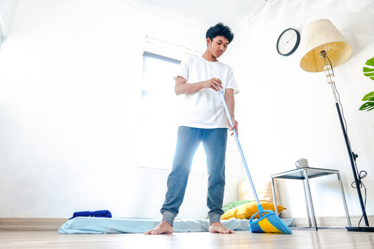 Young Asian Man Sweeping The Wooden Floor With Broom In Bedroom