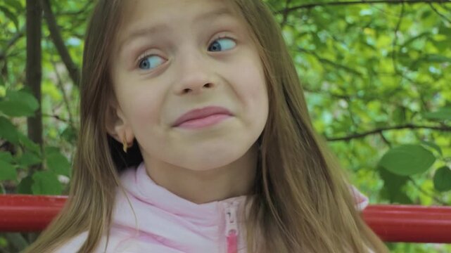 A joyful young girl makes a funny, cross-eyed face with wide blue eyes and hands on cheeks, capturing playful childhood humor against a green background.