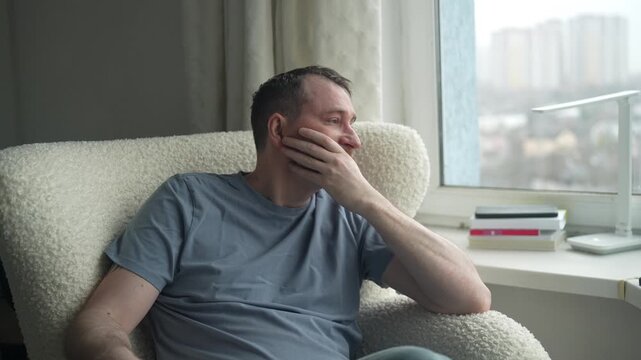 A man sits in a comfortable chair, deep in thought as he gazes out the window. The cloudy sky and city view create a calm atmosphere for reflection.