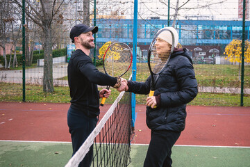 Man and woman shaking hands over a tennis net, symbolizing sportsmanship on an outdoor court. Autumn season.