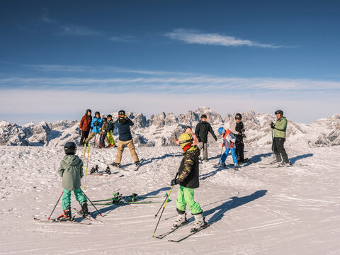 A group of people, including children, are skiing on a snowy mountain top under a blue sky. Paganella, Roda refuge,Trentino, Italy