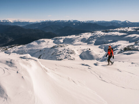 A woman snowshoes across a snowy mountain landscape under a clear blue sky. Paganella, Roda refuge,Trentino, Italy