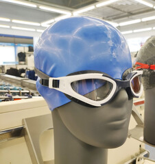 Mannequin head display of blue swimming cap and white swimming goggles in a retail store setting, showcasing essential swimwear accessories for sport and recreation