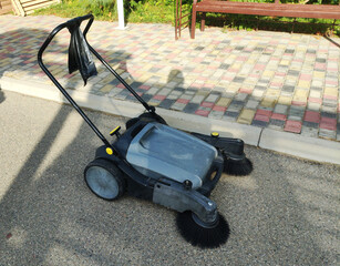 Manual push sweeper machine for outdoor floor cleaning, gray body and black handle, positioned on asphalt path near decorative paving stones and green garden area with wooden bench under daylight