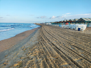 Ocean waves against shell fragments shoreline of beach with freshly raked sand and rows of beach cabins under clear blue sky morning light