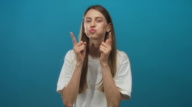 Young caucasian woman puffing cheeks and pointing fingers toward camera in teal studio with white tee visible; playful mischief.