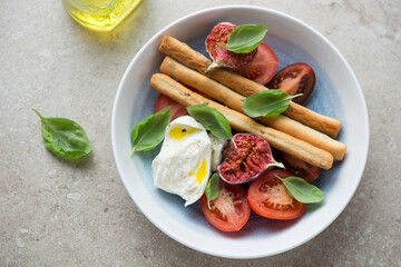 Tomato salad with fresh torn figs and mozzarella in a white and blue plate, horizontal shot on a beige stone background, high angle view