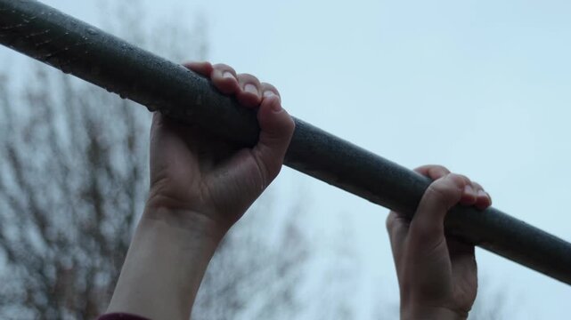 Fixed close-up of hands on a wet pull-up bar under a cloudy winter sky.